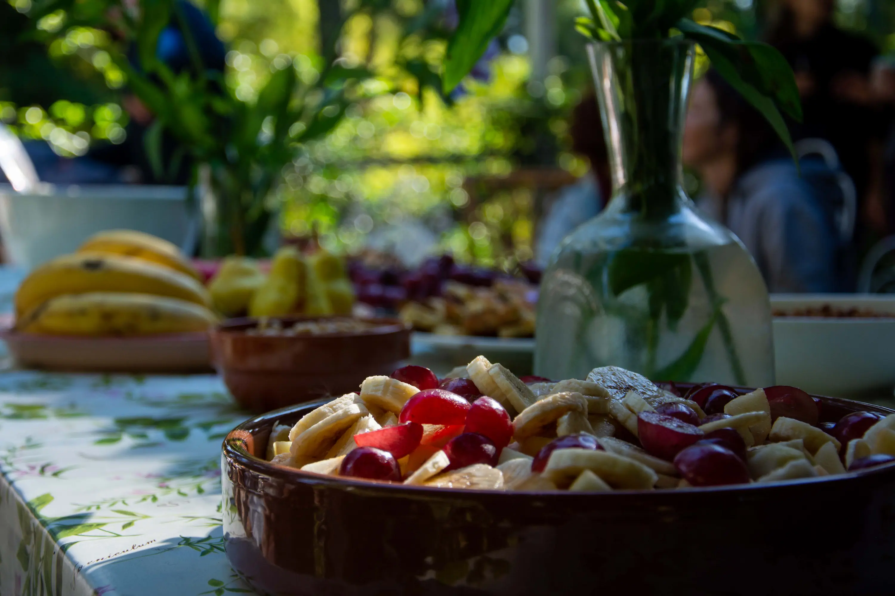 Una fuente de frutas cortadas sobre una mesa compartida al aire libre, con plantas de fondo
